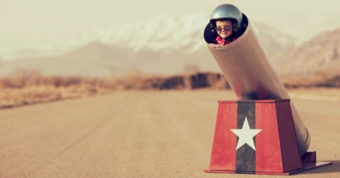 leadership conversations - A young child with light skin wearing a large, dark, shiny helmet and sunglasses is stuffed into a barrel of a pretend cannon propped up by a red blog with a navy vertical strip and large white star in the middle. The cannon is in the middle of an empty plane runway with mountains in the background.