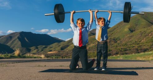 two children dressed in business clothes with ties holding up a big barbell above their heads with green mountains in the background.