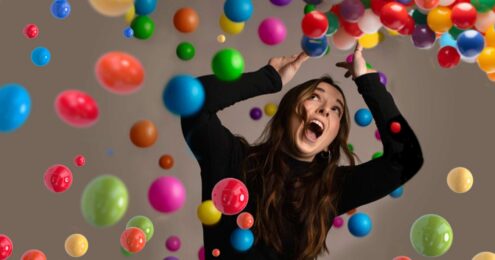 Image of a woman in a black long sleeve holding her head above her arms with a shocked look on her face as multi-colored balls fall down on her. Suggests the need for better time management.