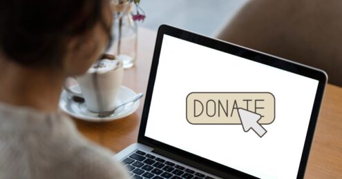 A person sitting at a wooden table in a café looks at a laptop screen displaying a large "DONATE" button with a cursor icon hovering over it. A coffee cup with frothy milk and a spoon rests beside the laptop, suggesting a relaxed environment for online giving.
