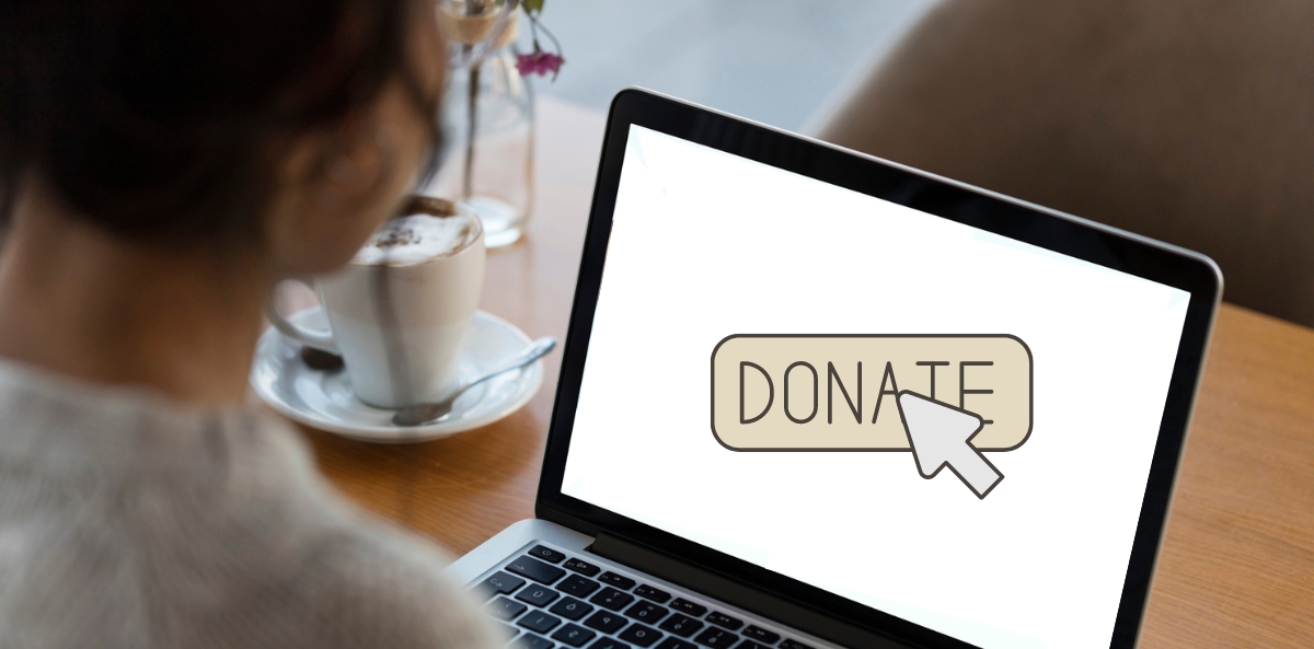 A person sitting at a wooden table in a café looks at a laptop screen displaying a large "DONATE" button with a cursor icon hovering over it. A coffee cup with frothy milk and a spoon rests beside the laptop, suggesting a relaxed environment for online giving.