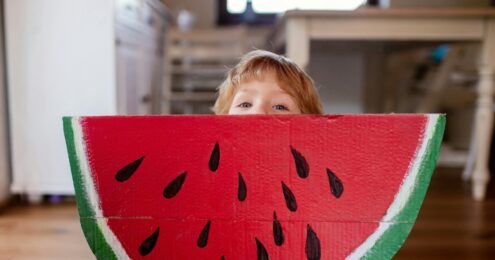 A child with an oversized slice of watermelon bigger than he is peers over the top.