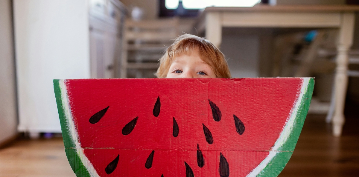 A child with an oversized slice of watermelon bigger than he is peers over the top.