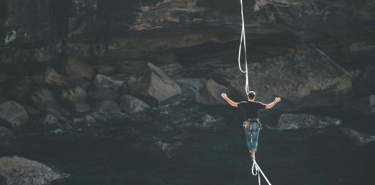 A person wearing a harness and climbing gear carefully walks across a highline stretched above dark, rocky waters, with arms extended for balance. A safety rope hangs overhead, and a towering cliff face looms in the background, emphasizing the height and risk of the challenge.