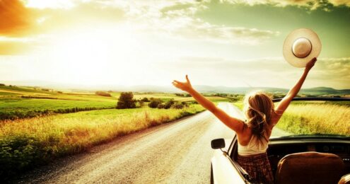 A woman with long blonde hair sits in a convertible car on a rural dirt road, facing a vast green landscape under a glowing sunset sky. She raises her arms joyfully, holding a wide-brimmed hat in one hand, embodying freedom and exhilaration on a summer drive through the countryside.