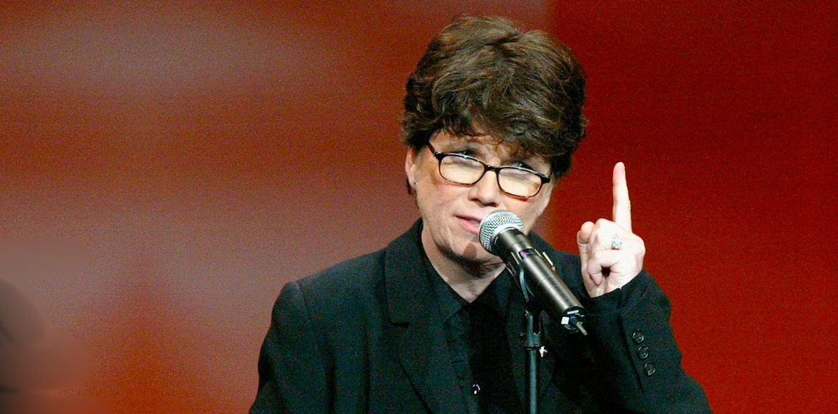 Joan Garry, age 39, stands confidently at a microphone against a warm red background. She wears a black suit and glasses, raising one finger in a thoughtful and expressive gesture as she speaks, conveying passion and conviction.