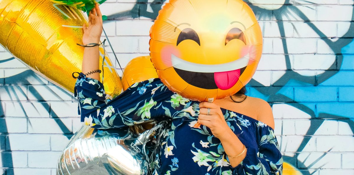A person in a floral top and jeans stands in front of a colorful mural, holding a smiley-face emoji balloon that covers their face. The scene reflects the pressure leaders often feel to appear cheerful even when they are overwhelmed.