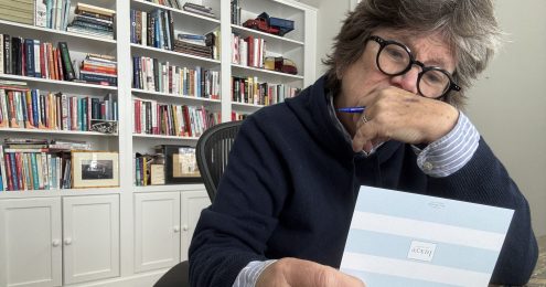 Joan Garry sits at her desk in a home office, holding a handwritten thank you appreciation note and pausing thoughtfully, with bookshelves behind her.