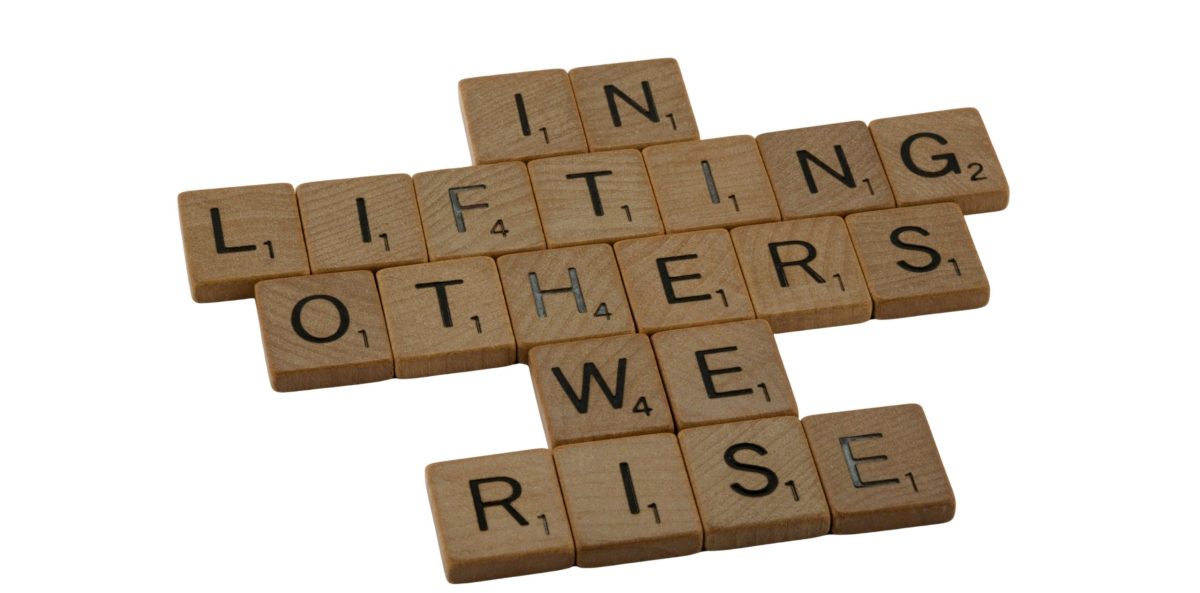 Wooden letter tiles arranged to spell the phrase “In lifting others we rise” on a light background.